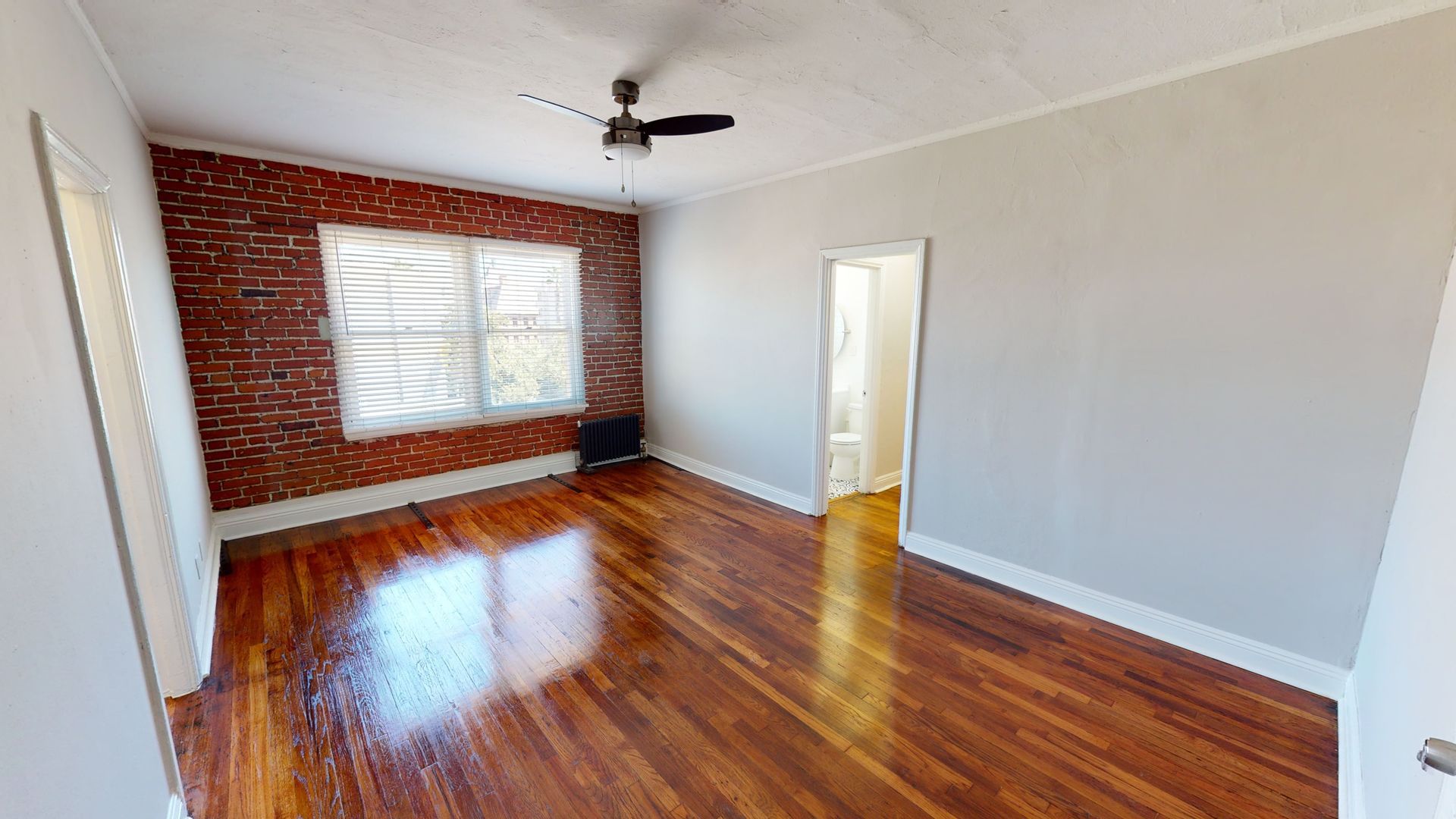 Room with brick wall and hardwood floor, window, doorway to bathroom, ceiling fan.