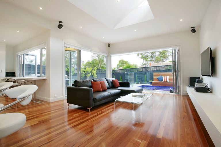 Bright modern living room with hardwood floors, dark sofa, white chairs, and large windows opening to a pool. — KM Custom Homes In Mount Eliza, VIC