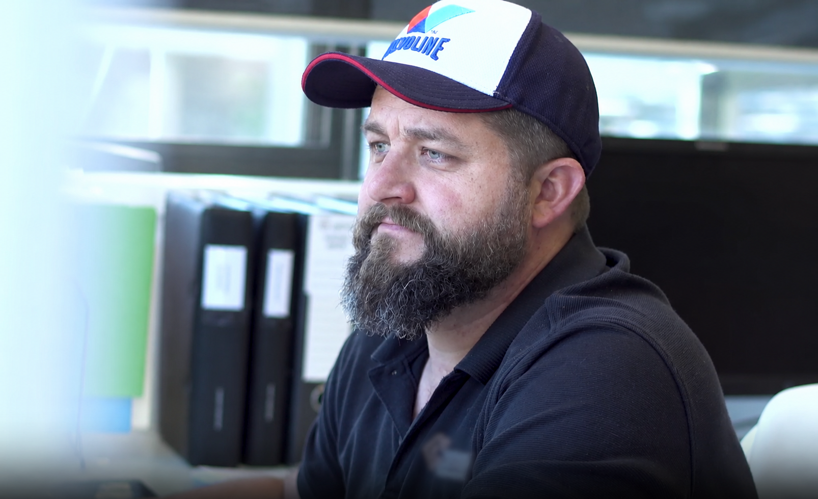 Bearded man in a dark shirt and cap sitting at a desk in an office, looking to the side. — KM Custom Homes In Mount Eliza, VIC