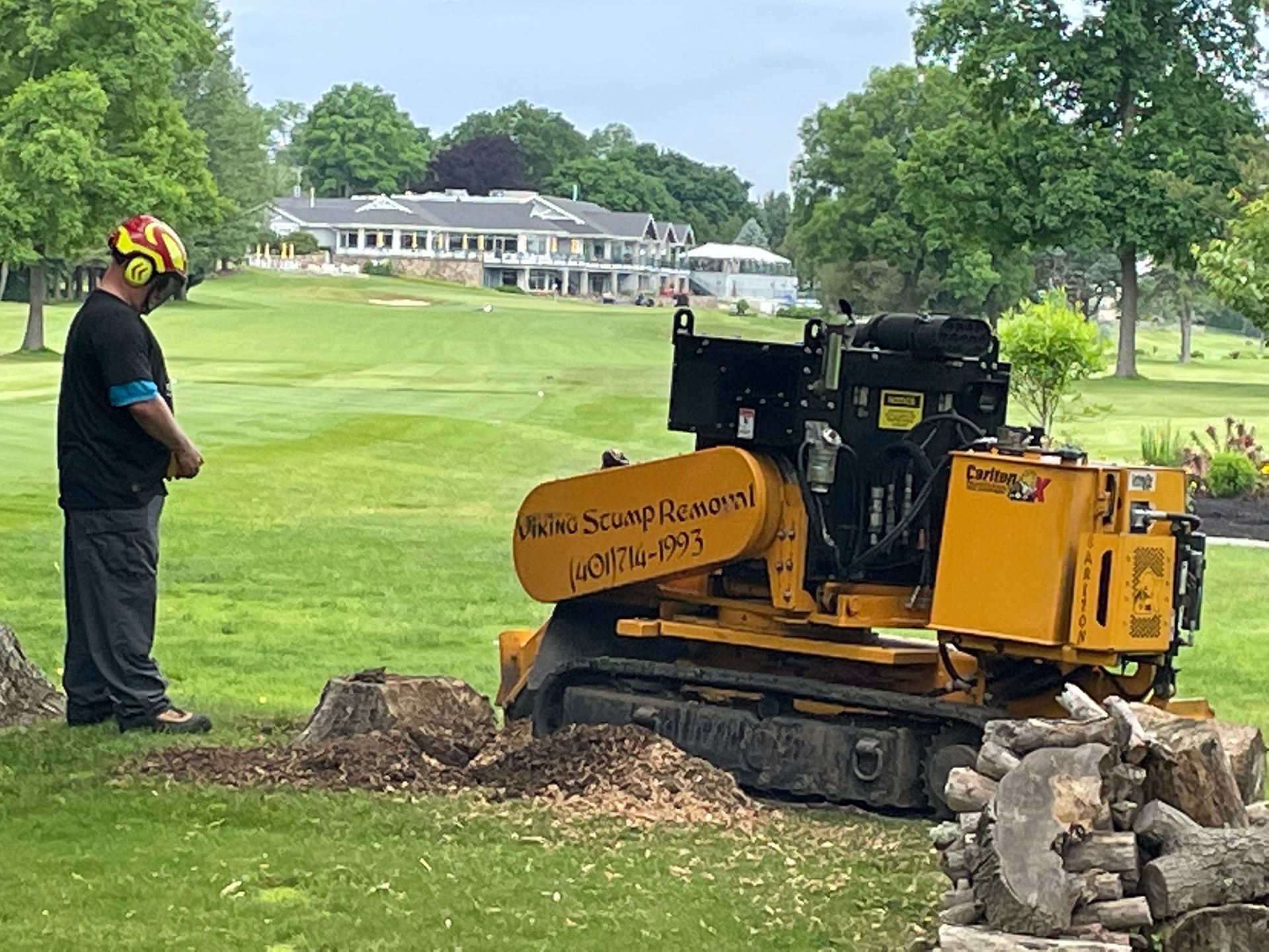 Worker with helmet operates a yellow stump grinder beside rocks on a grassy lawn near a large house.