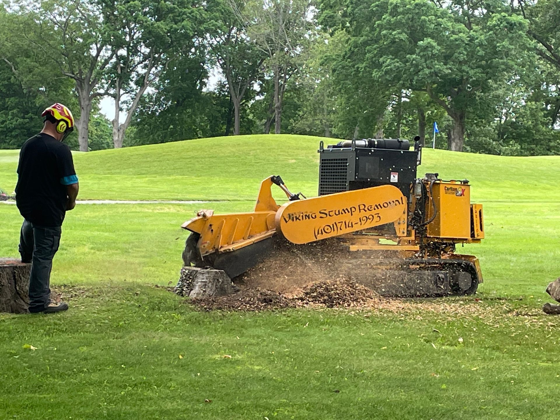 Worker stands beside a yellow track loader digging turf in a grassy park.