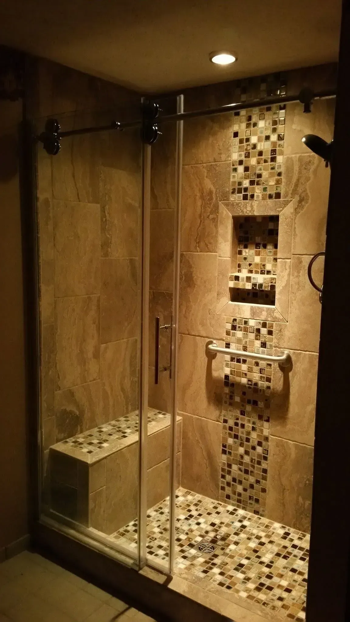 Glass-enclosed shower with mosaic tile walls and built-in bench, lit by warm ceiling lights