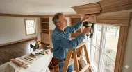 Worker installing trim around a window in a bright room, with tools on a table and a ladder nearby