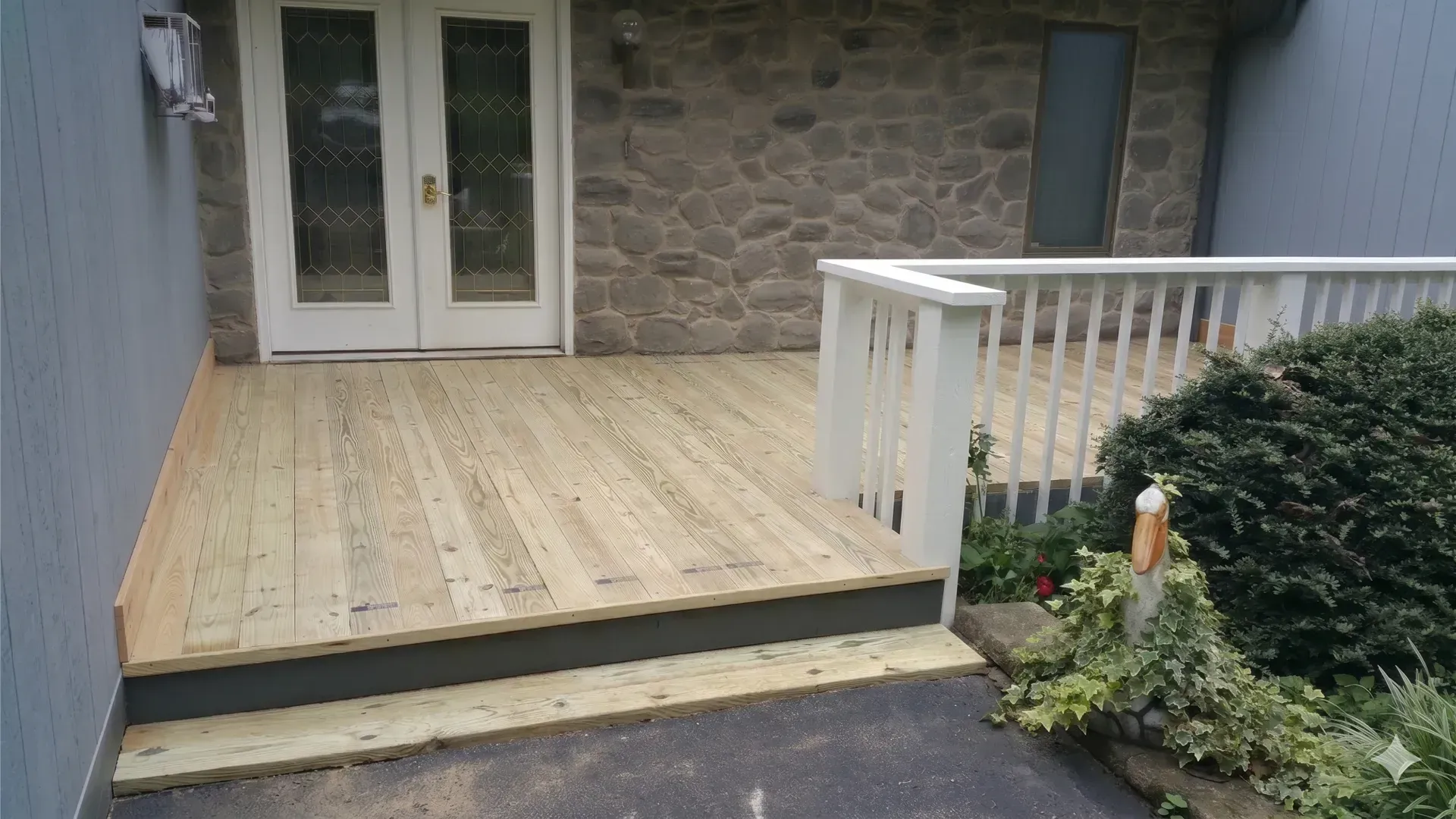 Wooden deck leading to white double doors beside a stone wall and white railing with shrubs.