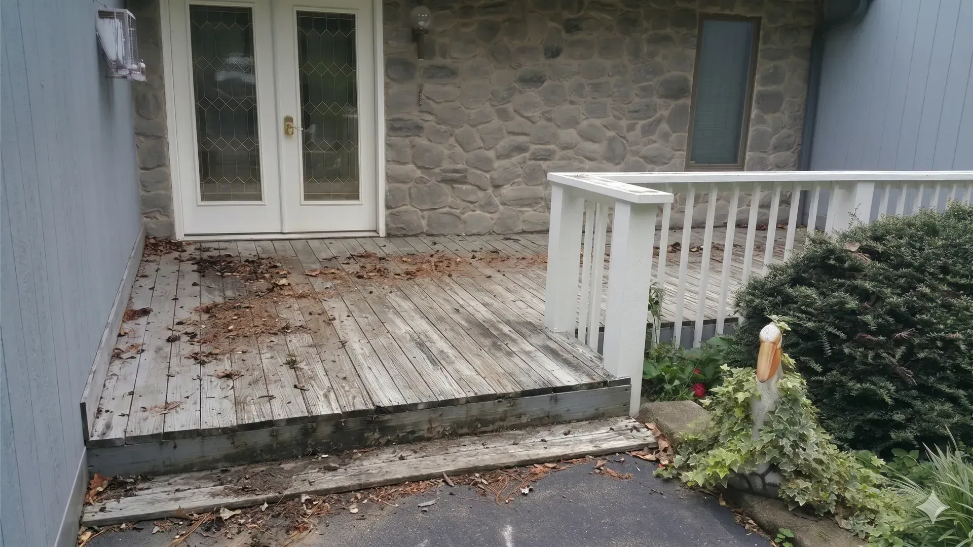 Weathered wooden deck with white railing outside a stone house, covered in scattered leaves.