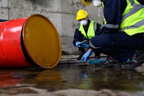 Two men wearing hard hats and safety vests are kneeling next to a barrel.