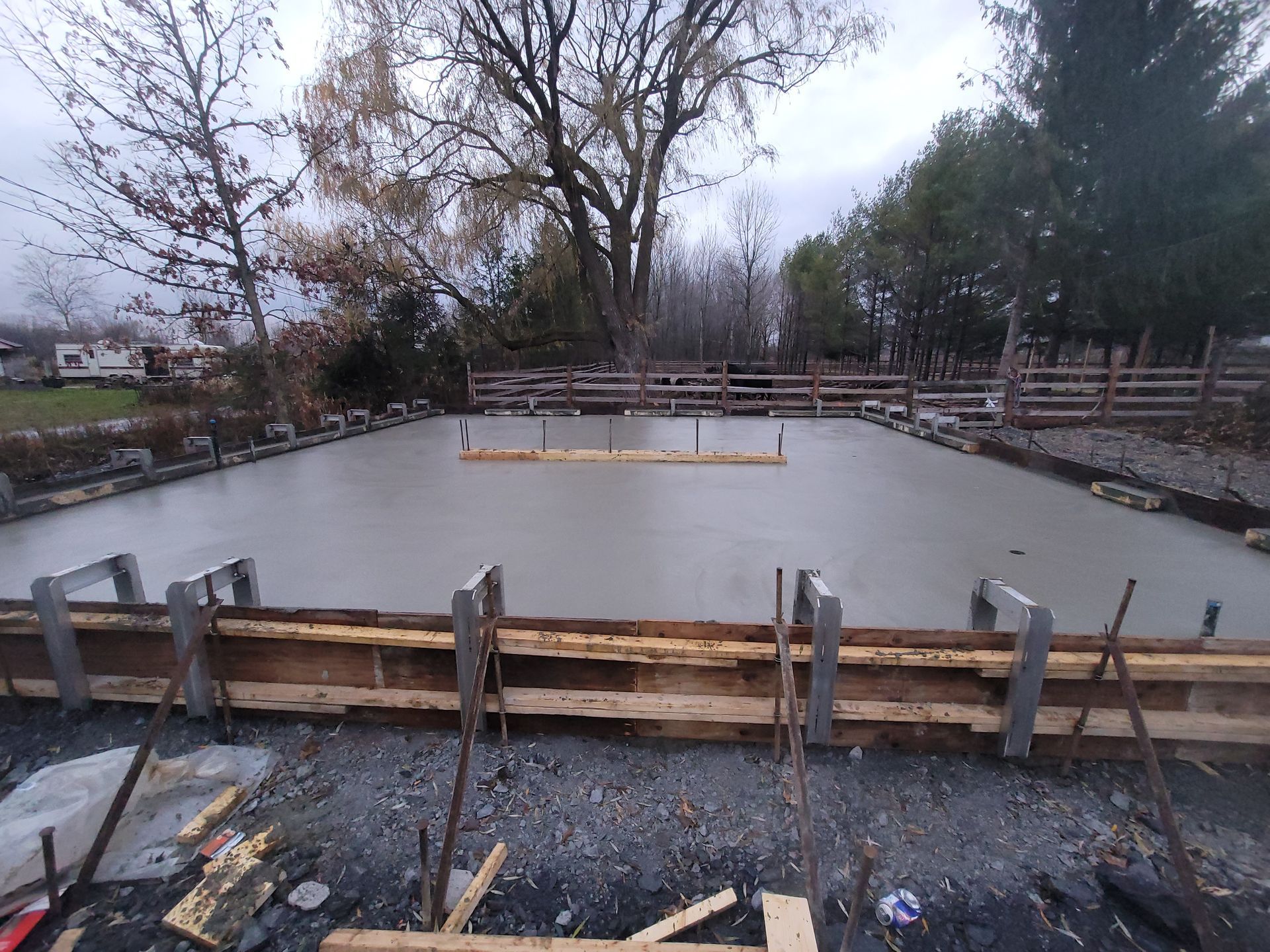 Fondations en béton fraîchement coulées dans des coffrages en bois sur un chantier de construction, entourées d'arbres et de clôtures.