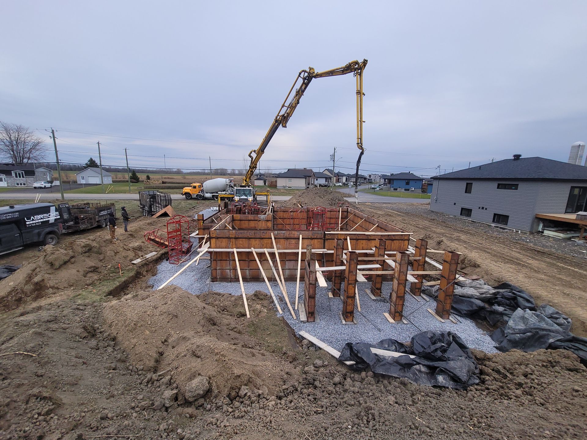 Chantier de construction avec une excavatrice coulant du béton dans des coffrages de fondation en bois sous un ciel nuageux