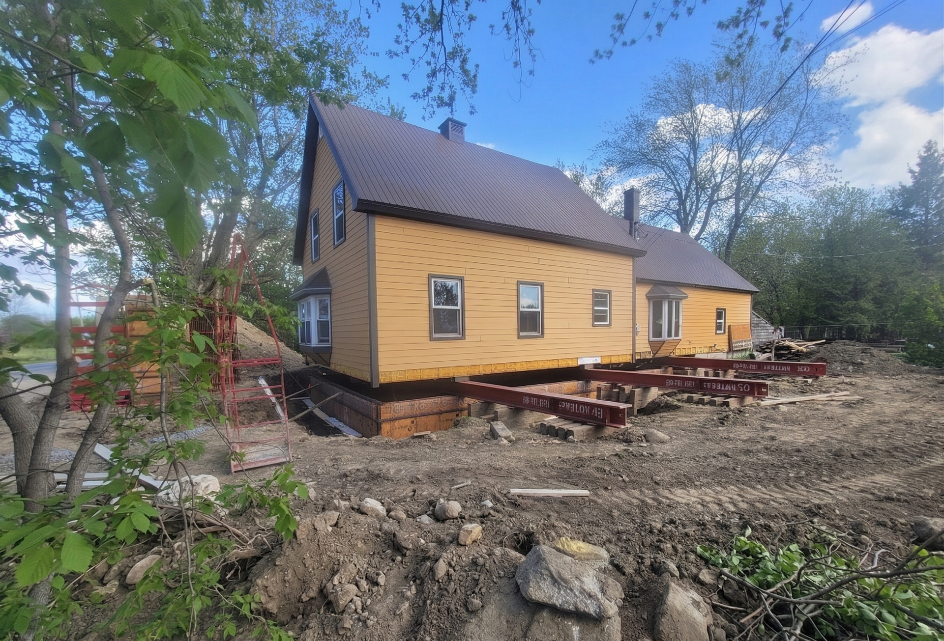 Maison jaune en construction sur fondations surélevées, entourée d'arbres et de terrain boueux.