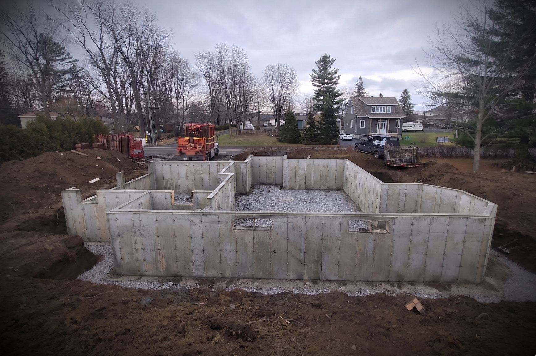 Fondations en béton pour une maison en construction sur un terrain boueux, avec une excavatrice et des maisons en arrière-plan.