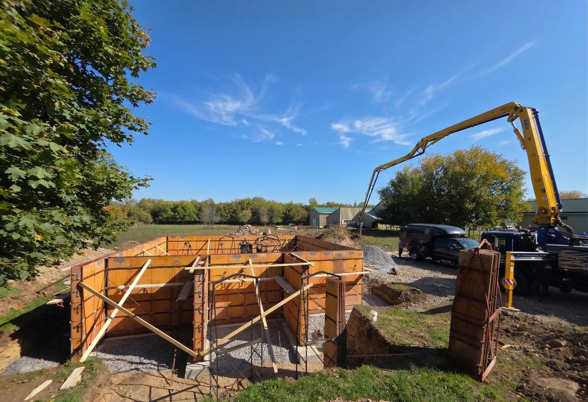 Chantier de construction avec coffrages de fondation en béton, excavatrice et véhicule stationné sous un ciel bleu dégagé