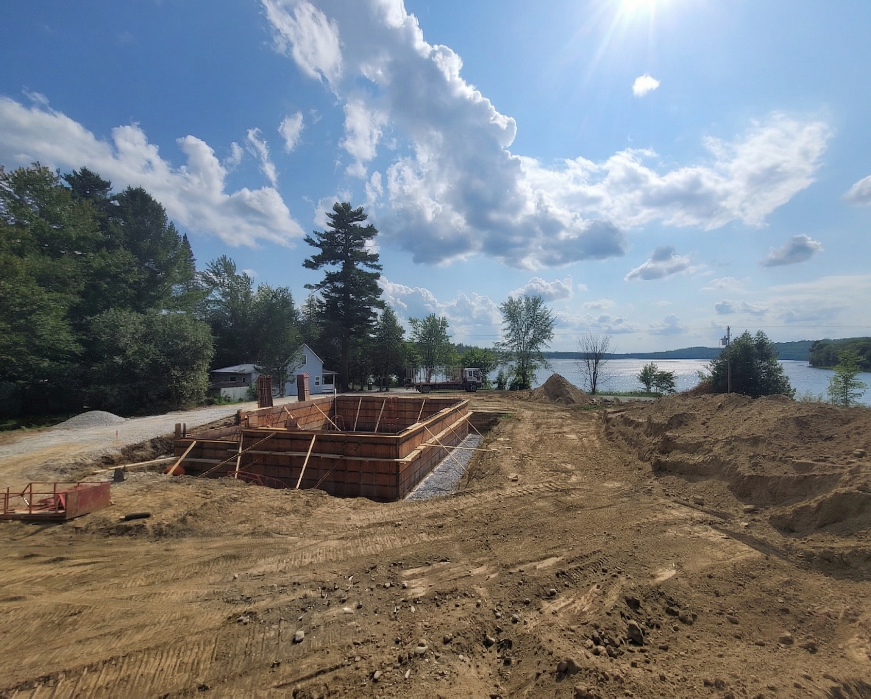 Chantier de construction avec coffrages de fondation en bois sur un terrain en terre battue surplombant un lac sous un ciel d'un bleu éclatant.
