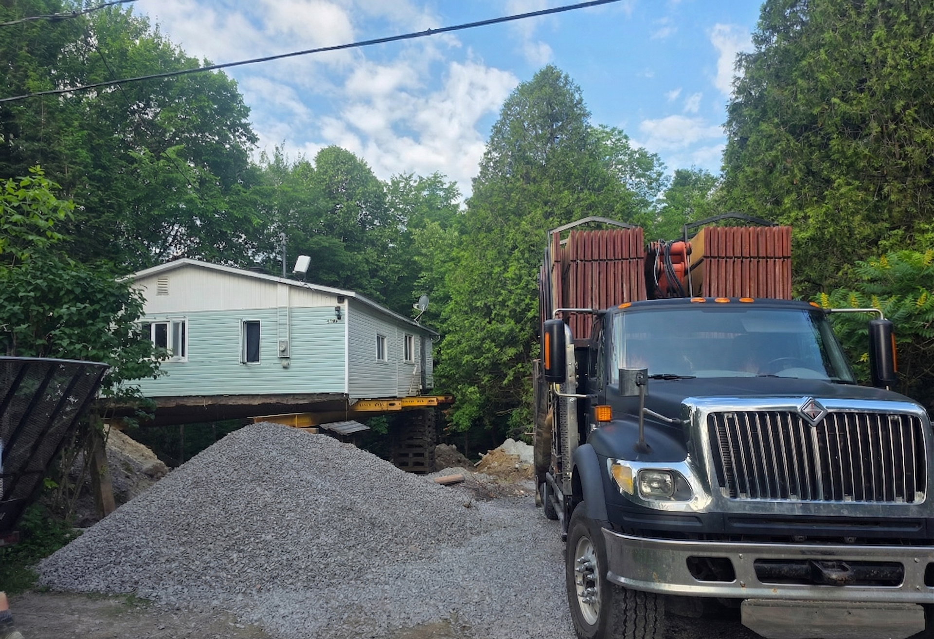 Maison soulevée par des crics à côté d'un camion-benne et d'un tas de gravier dans une cour boisée