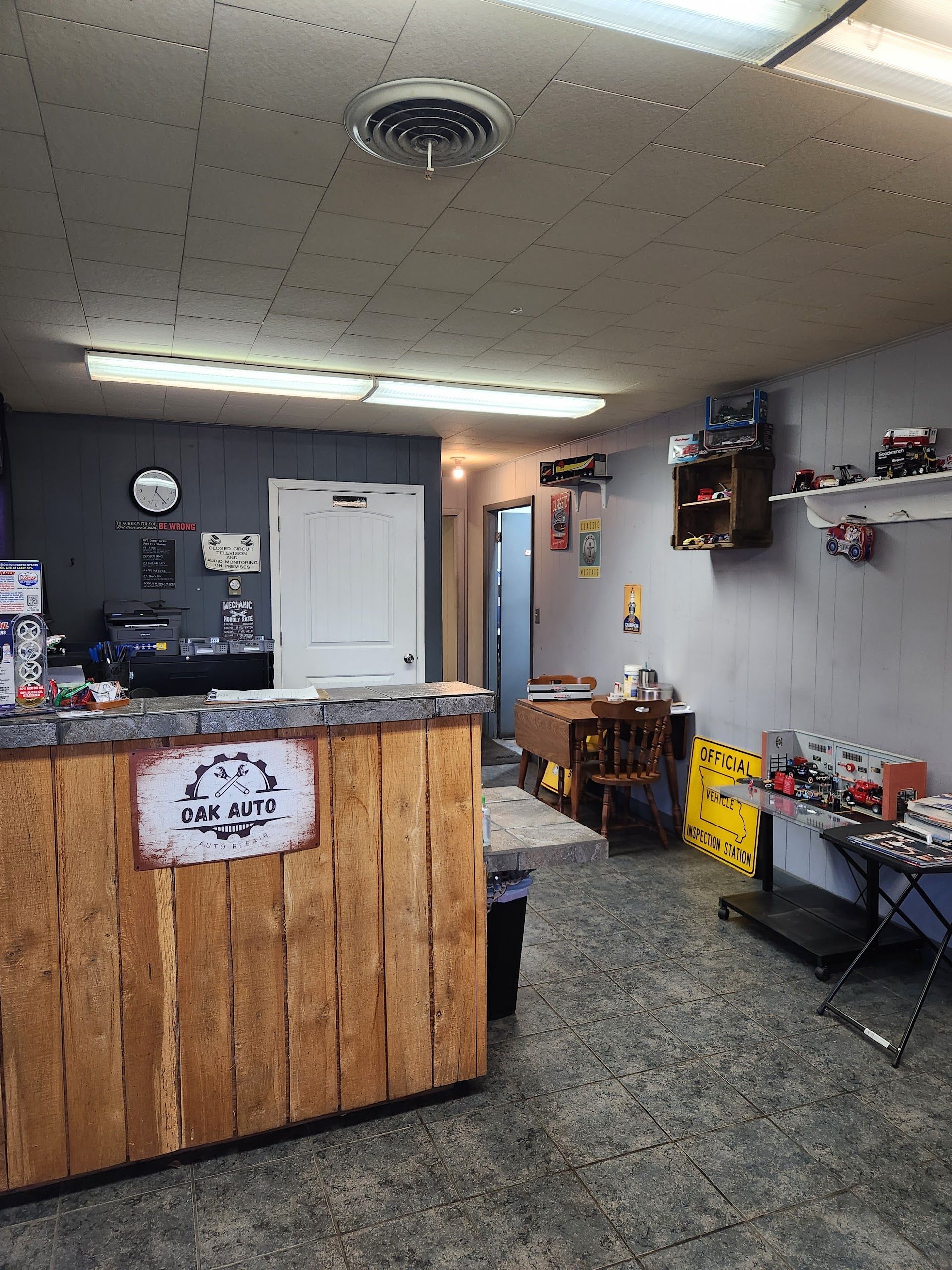 Small reception area with wooden front desk, white door, wall clock, shelves, and patterned floor. | Oak Auto