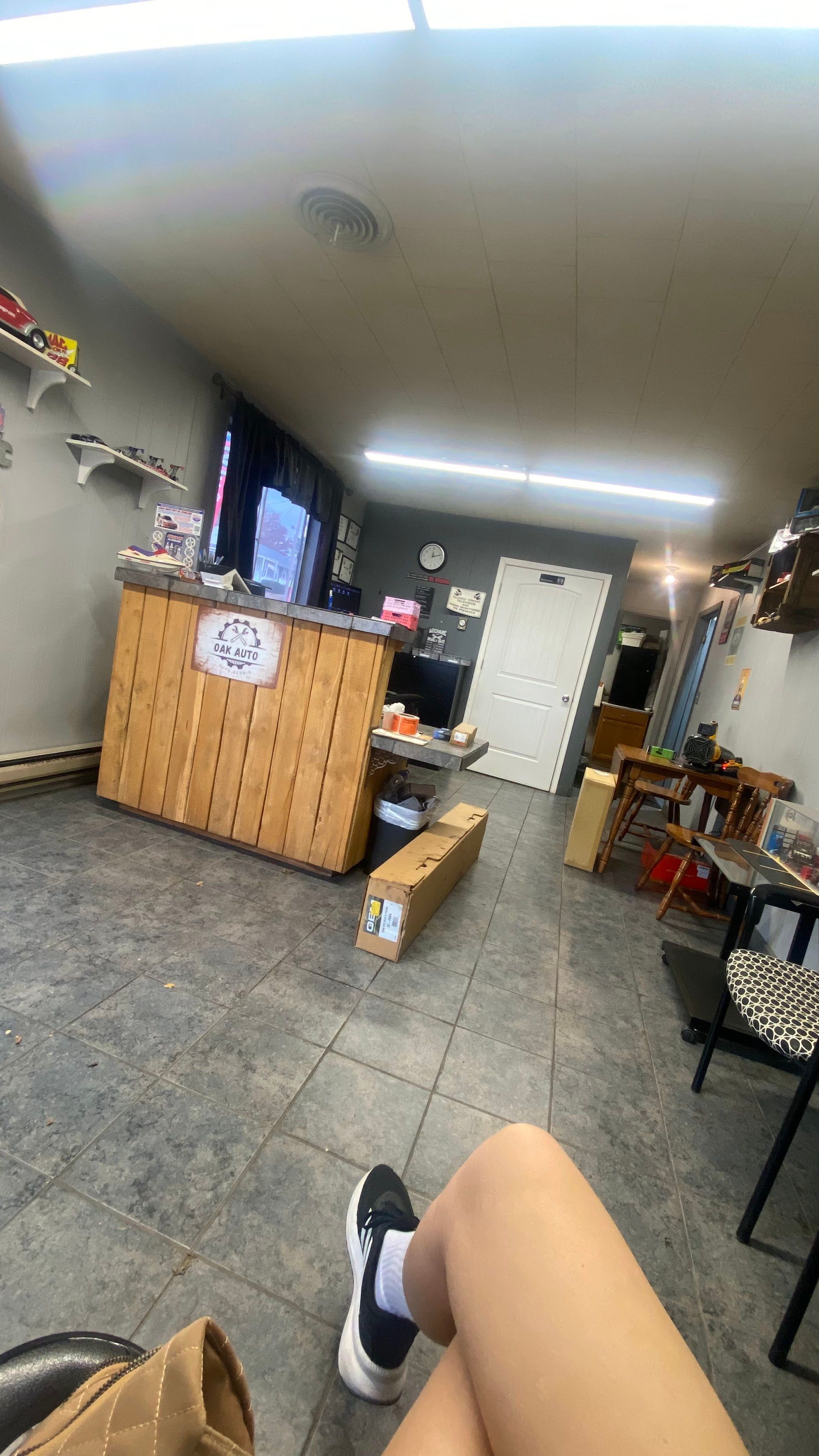 Tilted view of a cozy room with a wooden counter, wall clock, scattered boxes, and a seated person’s leg in foreground | Oak Auto