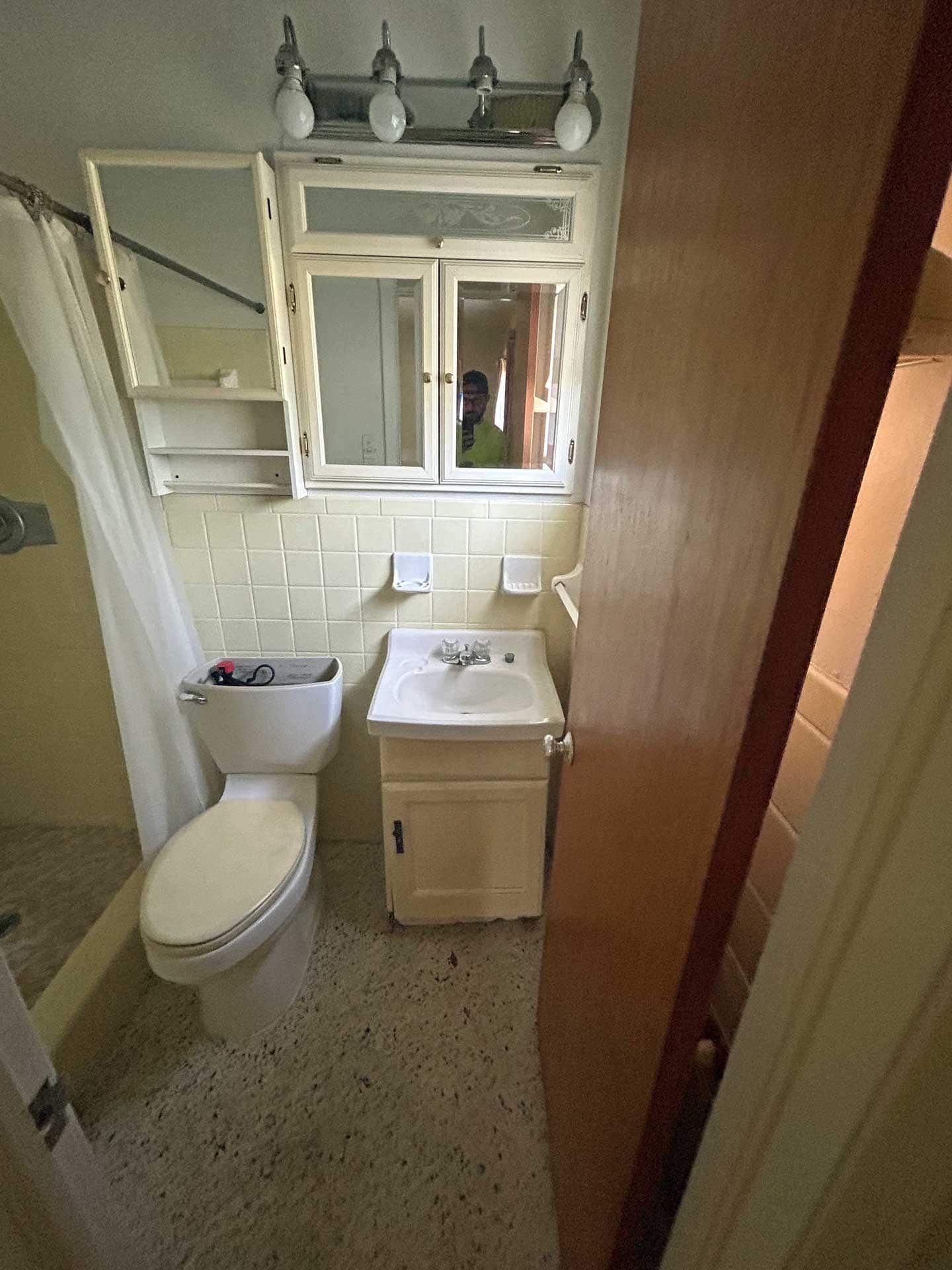 Small bathroom with a toilet, sink, and shower. Yellow walls and tile, light floor, and wooden door.