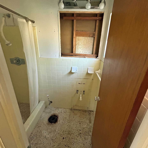 A bathroom with a shower, partially torn-out wall, and damaged flooring. Brown door on the right.