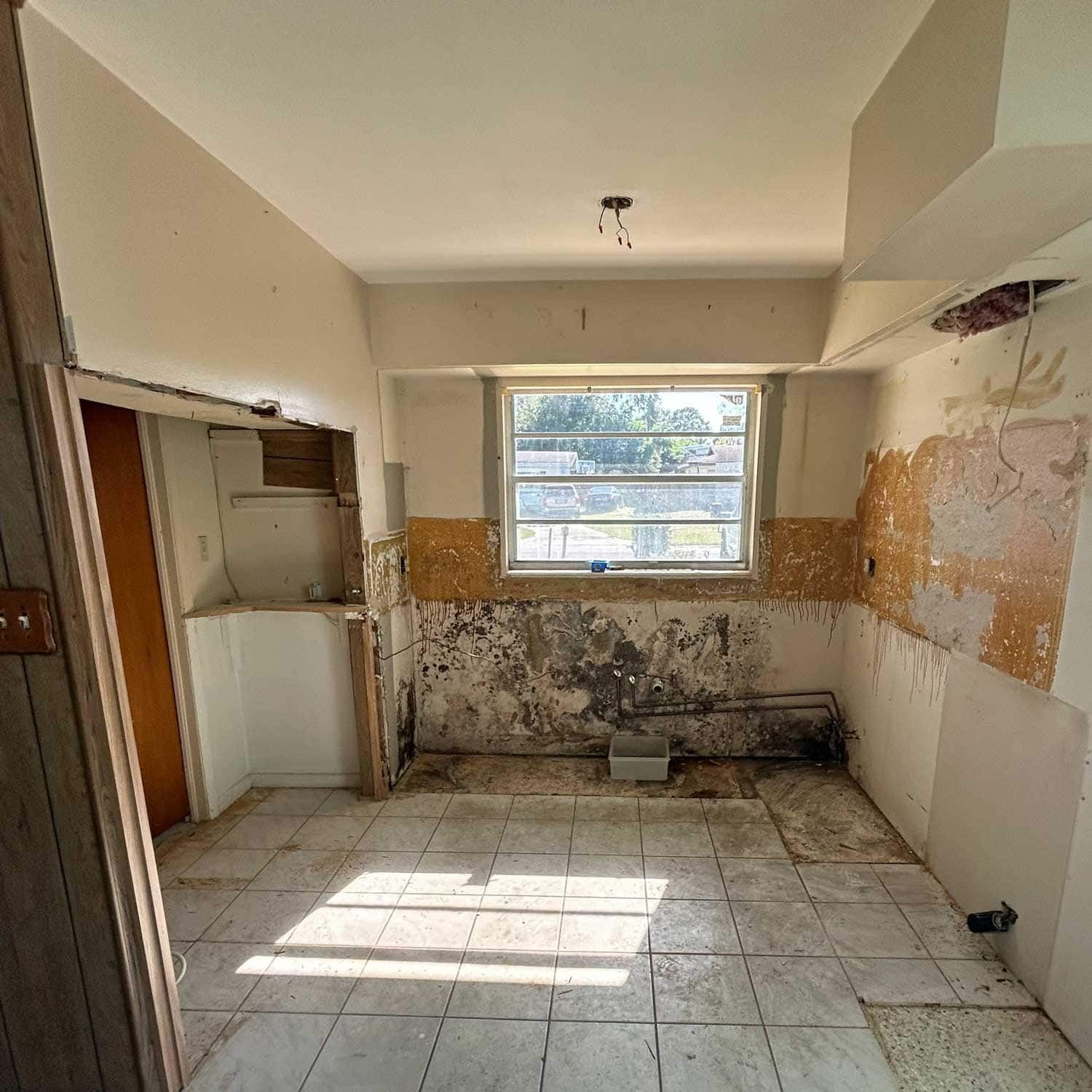 Kitchen undergoing renovation: walls stripped, cabinets removed, debris on floor, window visible.