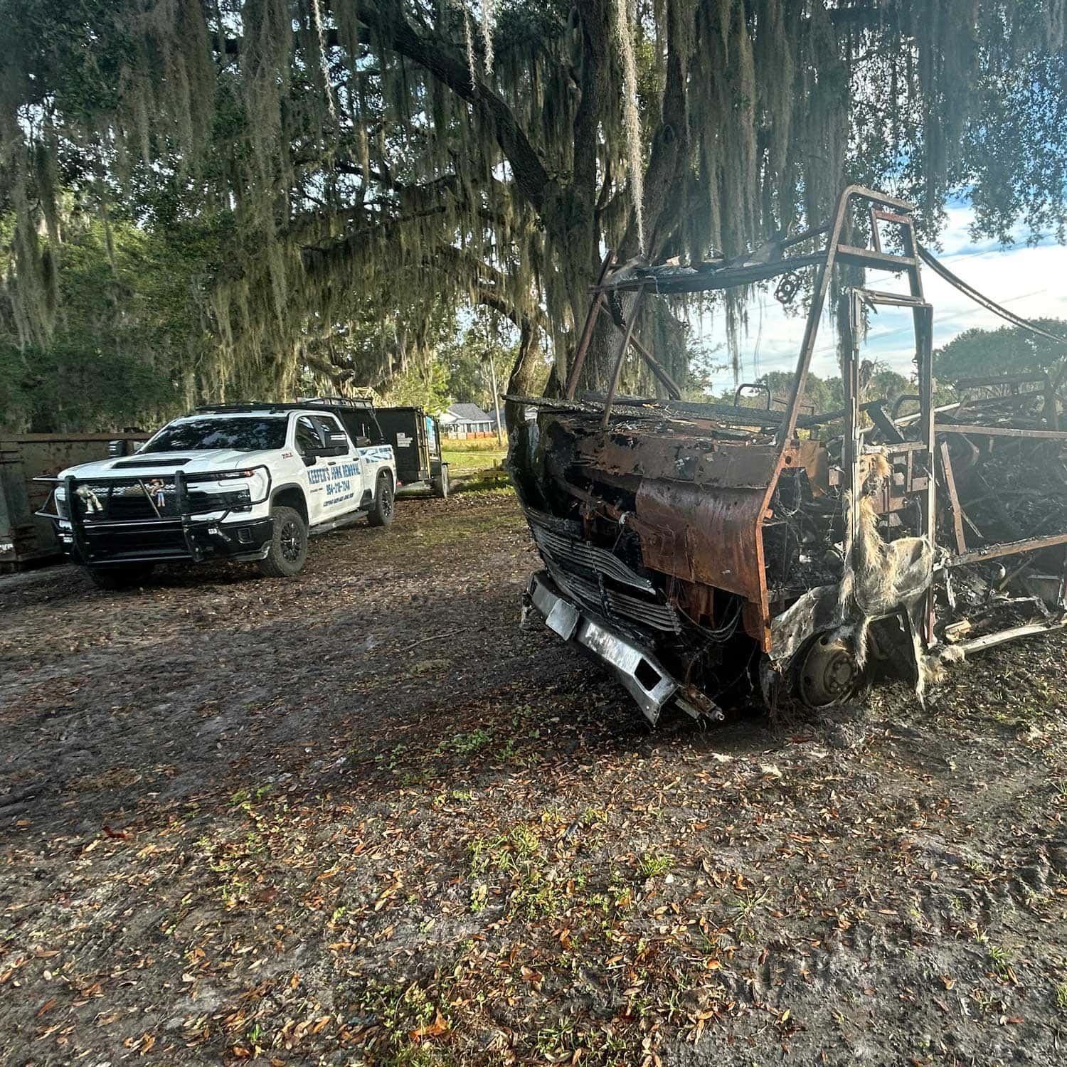 Burned-out vehicle beside a law enforcement truck in a wooded area. Debris on the ground, trees in the background.