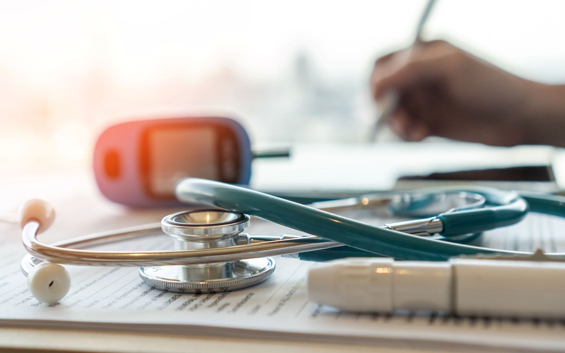 Stethoscope, glucose meter, and pen on paperwork; a hand writes in the background.