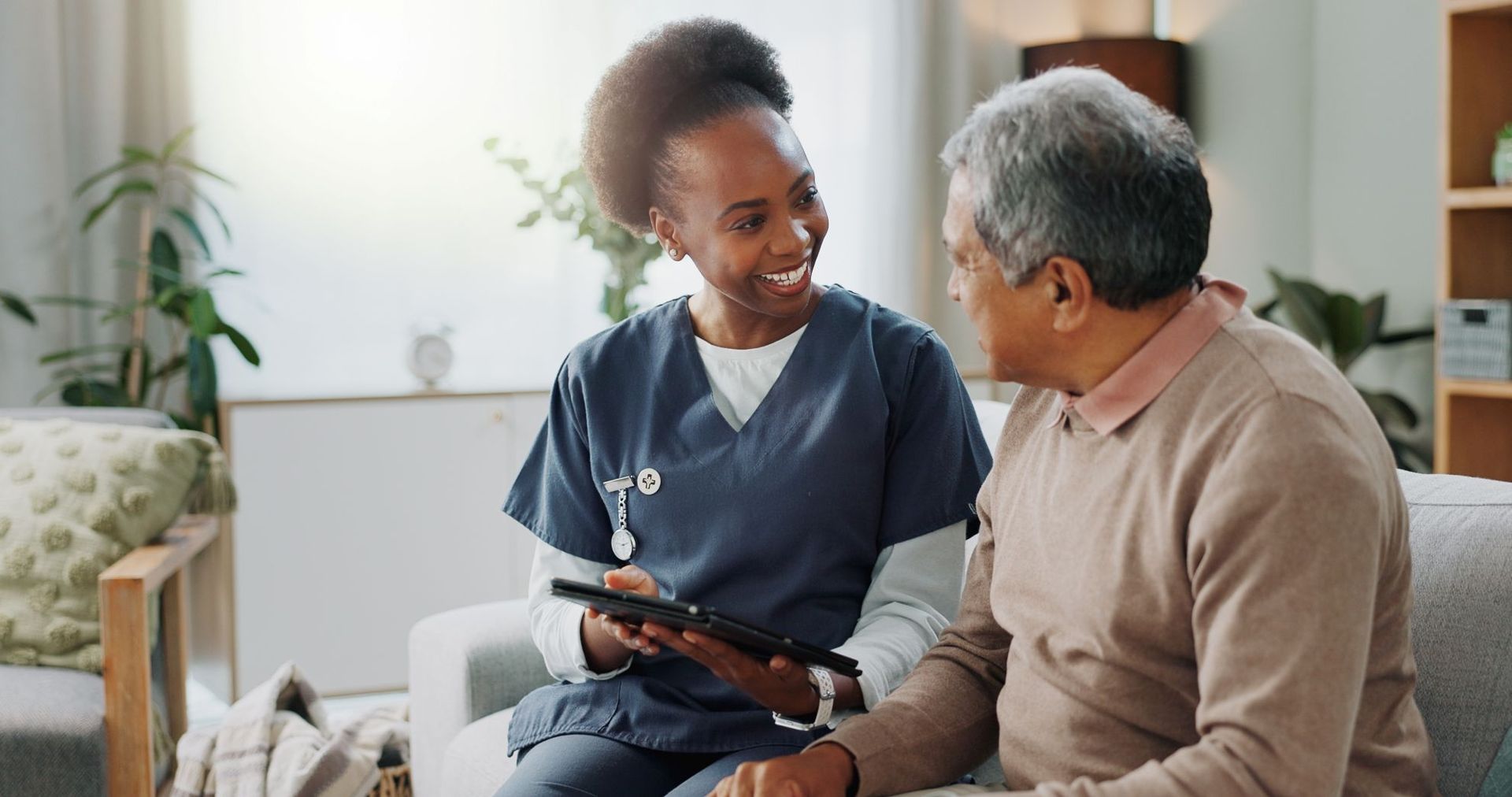 Nurse in blue scrubs showing tablet to seated patient on a couch indoors.