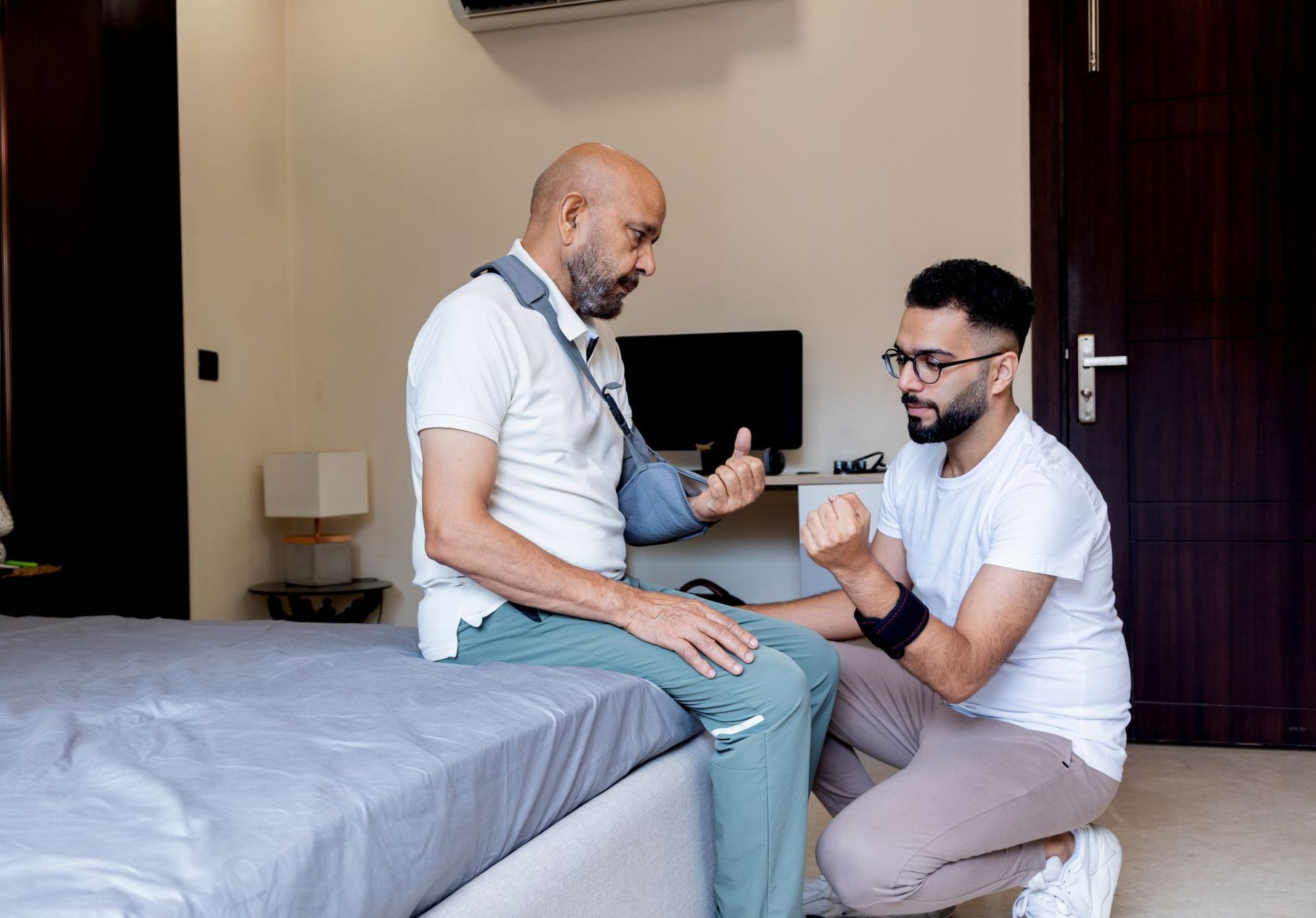 Man with arm sling on bed, interacting with a person kneeling, possibly providing assistance. Room setting.