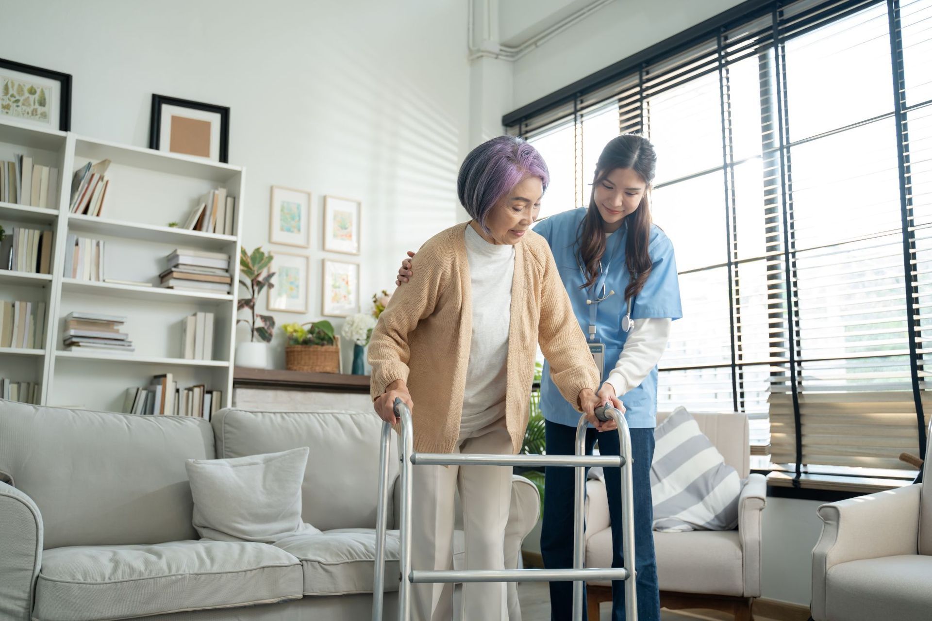 Elderly person using a walker, assisted by a healthcare worker indoors.
