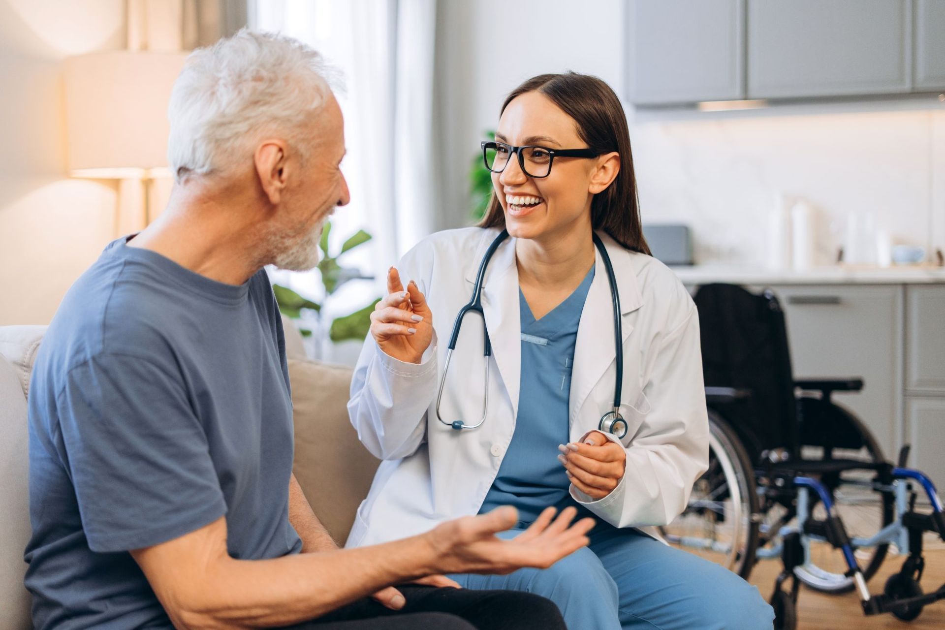A doctor in a white coat smiles, speaking with an older man seated on a sofa. A wheelchair is in the background.