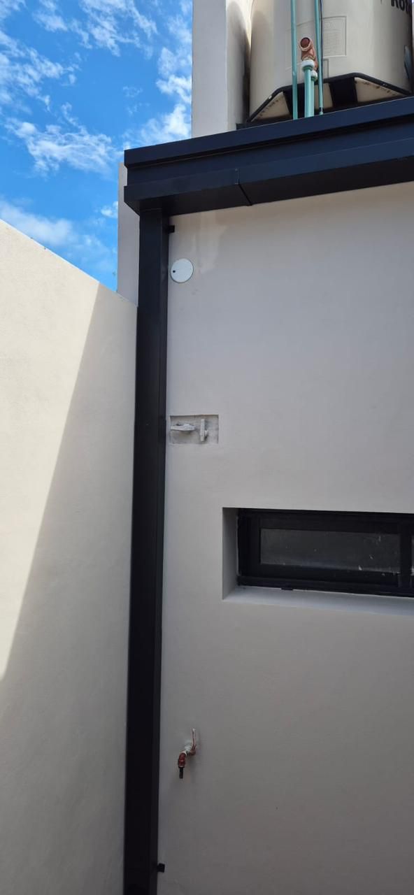 Pared exterior con una estrecha ventana negra, bajante y depósito de agua en la azotea, con un cielo azul de fondo.