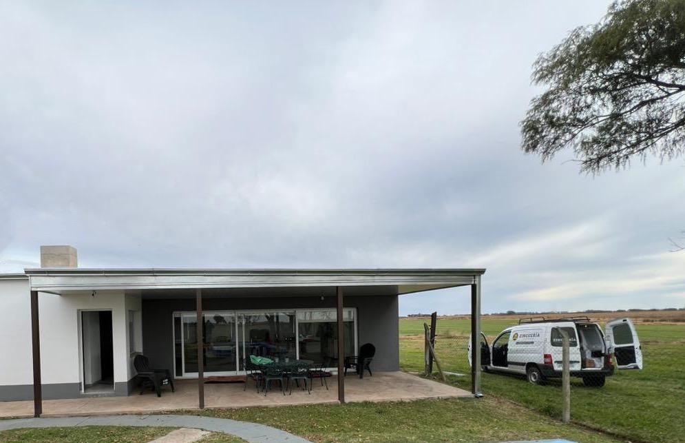 Pequeña casa moderna con porche cubierto, furgoneta blanca aparcada y cielo nublado en un terreno cubierto de césped.