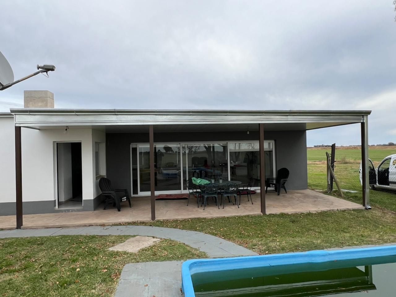 Casa blanca moderna con patio cubierto, mesa y sillas de exterior, y un jardín con césped junto a una pequeña piscina.