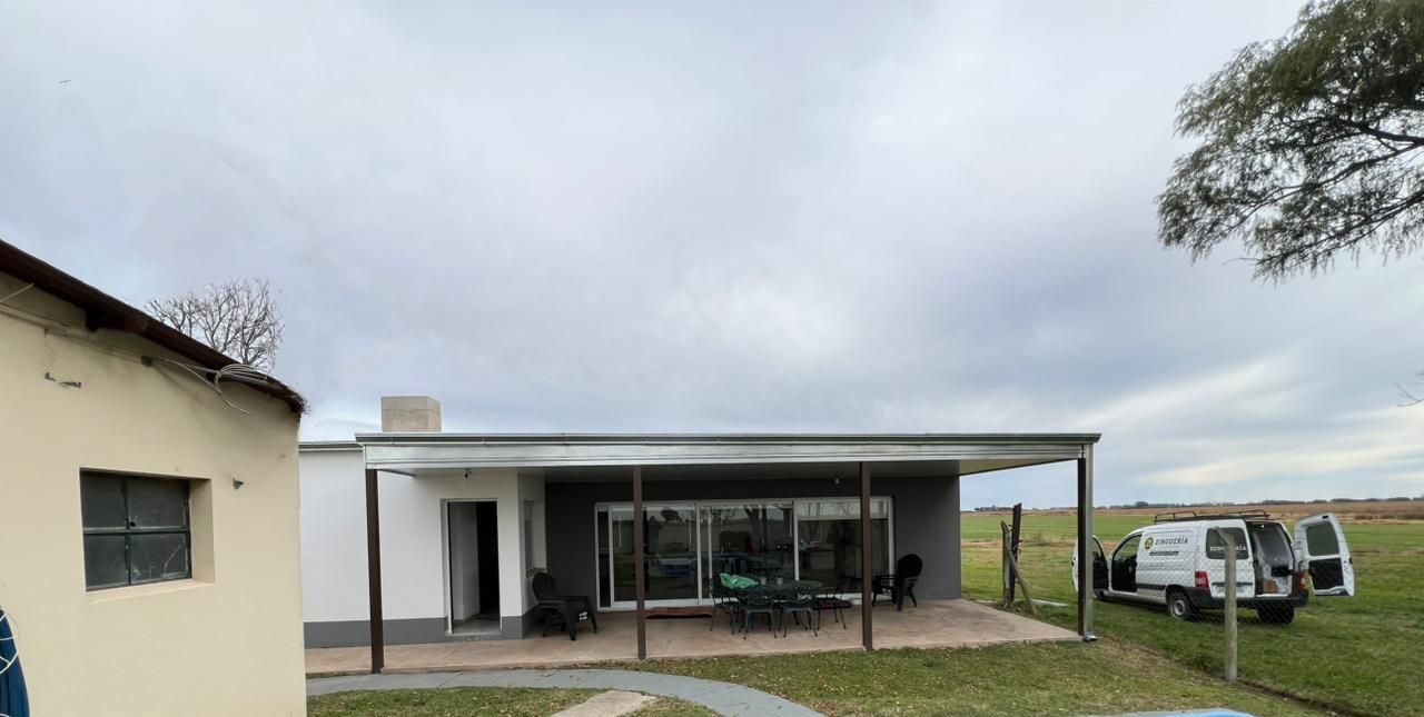 Pequeña casa con patio cubierto y furgonetas aparcadas en un terreno cubierto de hierba bajo un cielo nublado.