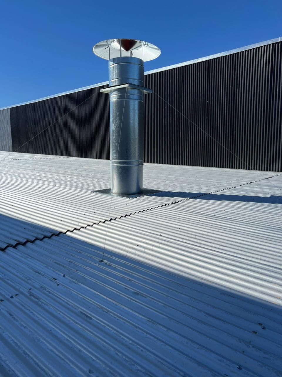 Tubo de ventilación metálico en un tejado ondulado junto a una pared negra bajo un cielo azul claro.
