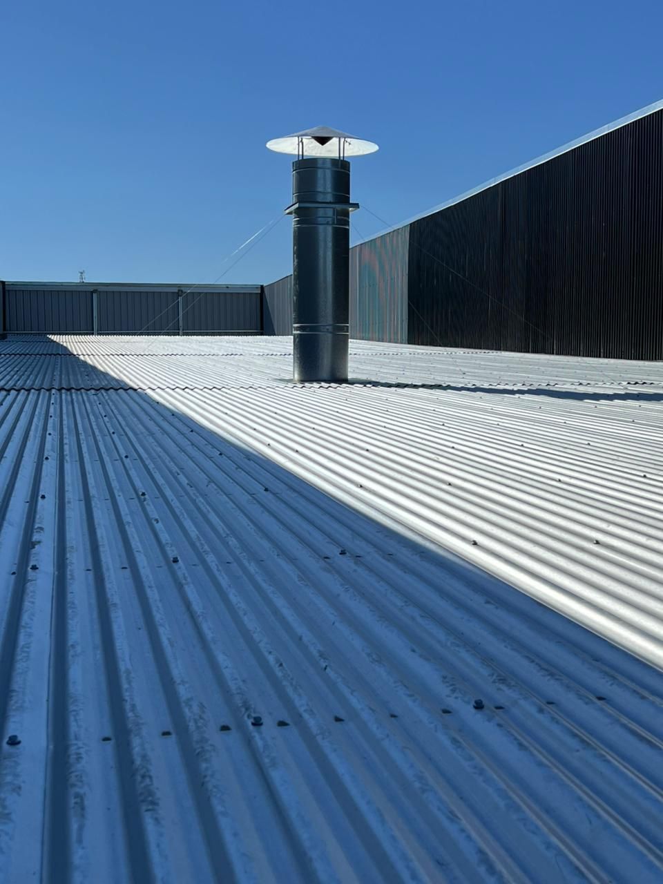 Techo de chapa ondulada con un tubo de ventilación negro bajo un cielo azul claro.