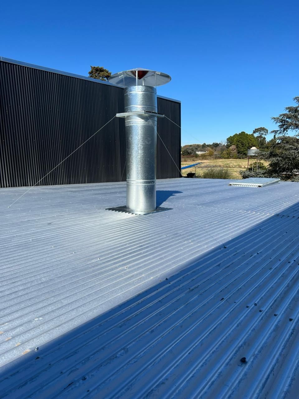 Tubo de ventilación de metal plateado sobre un tejado ondulado, junto a un edificio negro y un paisaje árido.