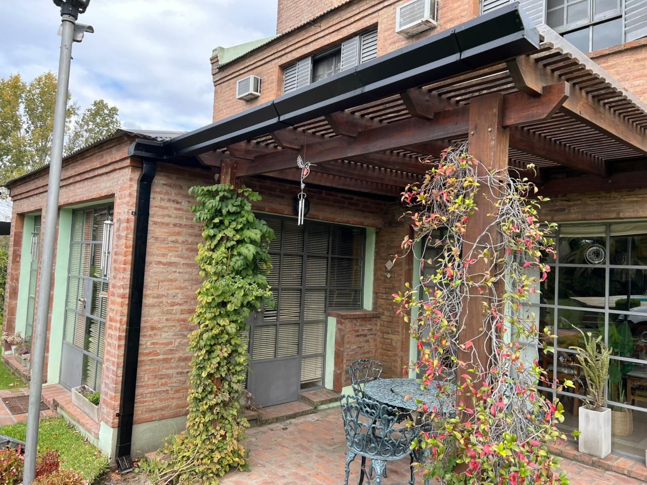 Patio de casa de ladrillo con pérgola de madera, plantas trepadoras y asientos al aire libre.