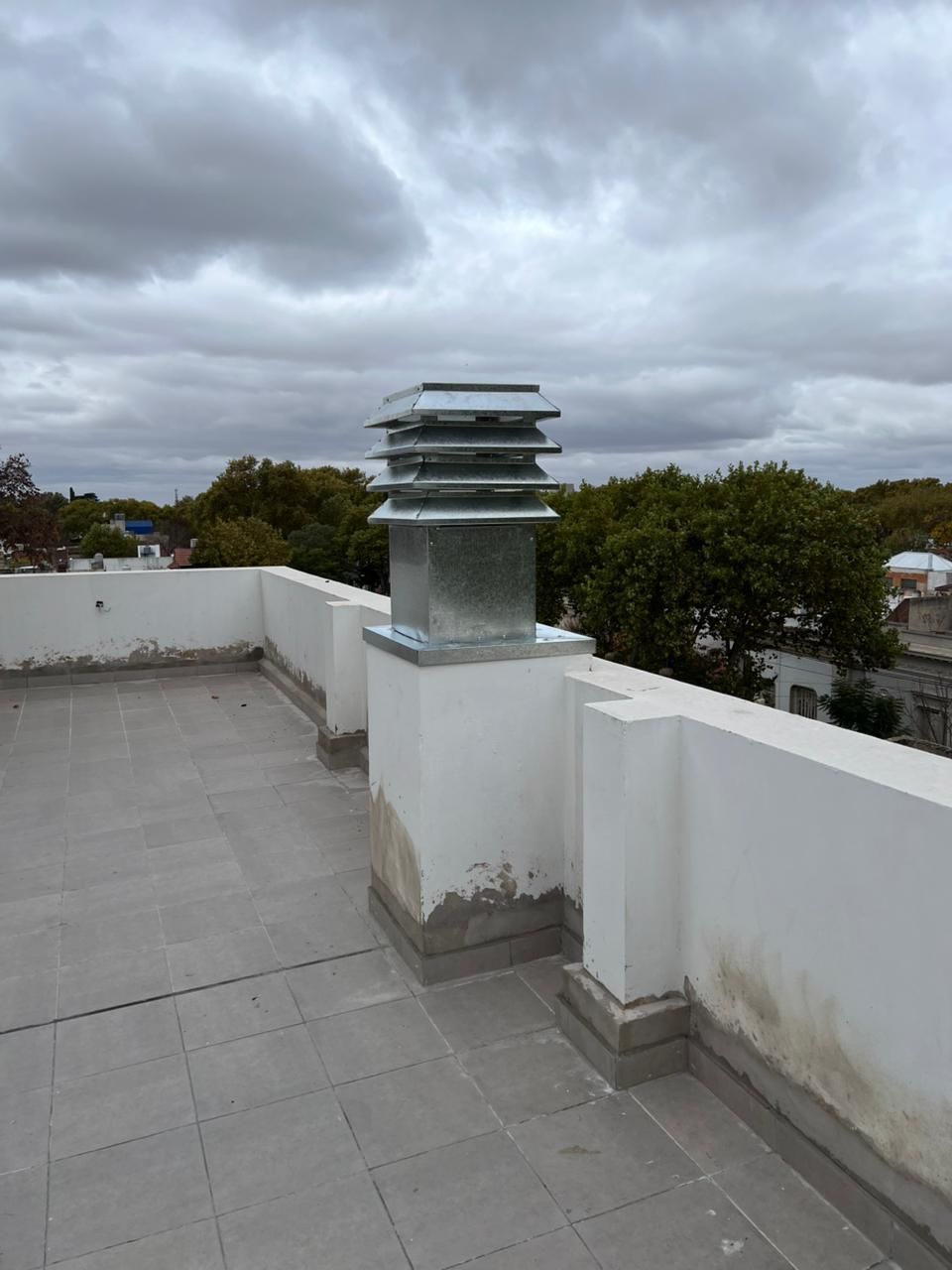 Terraza en la azotea con paredes blancas, azulejos grises y una rejilla de ventilación metálica bajo un cielo nublado.