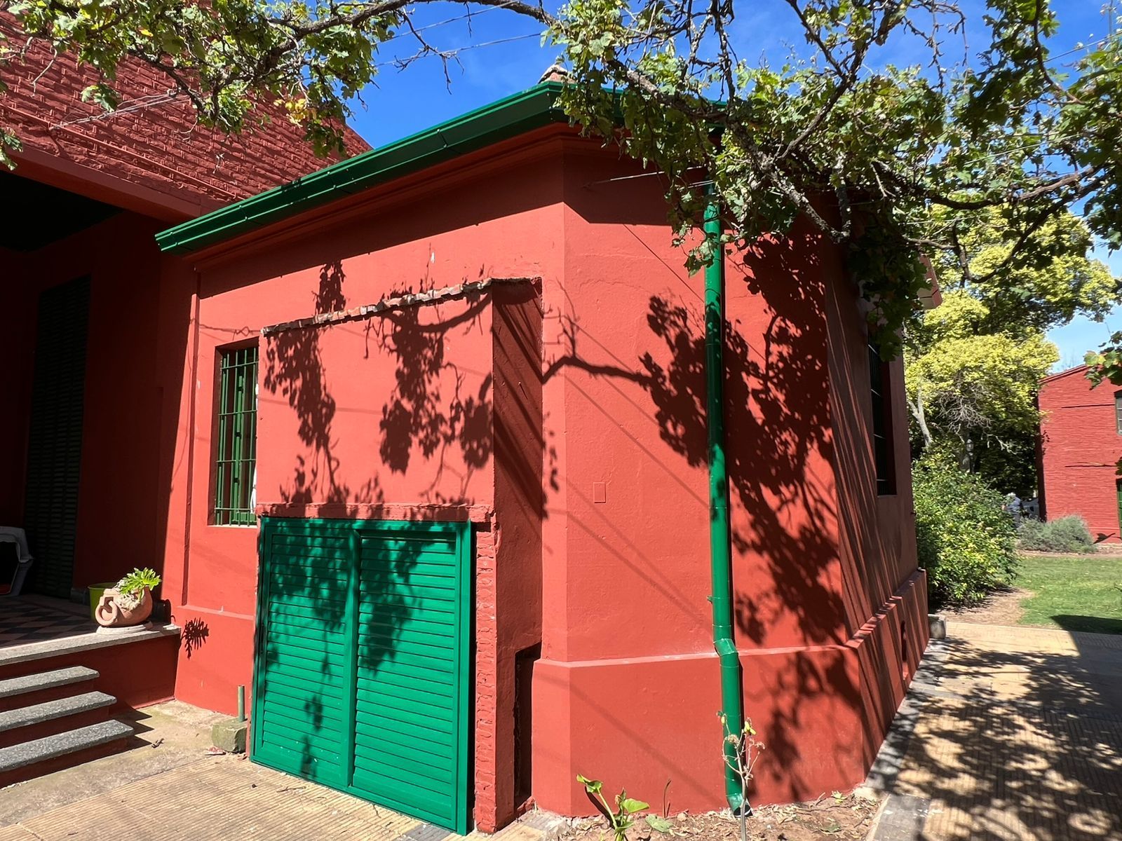 Edificio rojo con puertas y molduras verdes bajo los árboles, junto a escalones de piedra en un patio soleado.