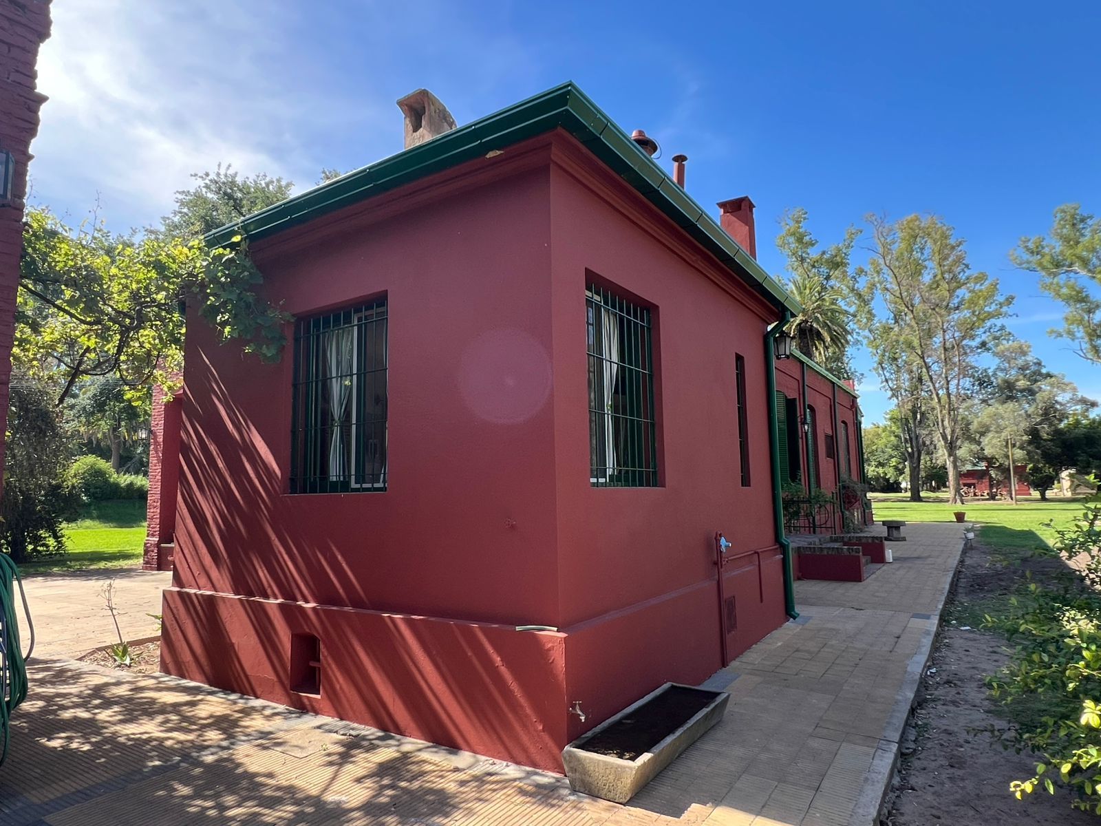 Edificio rojo con detalles verdes y ventanas enrejadas junto a una acera en un parque soleado.