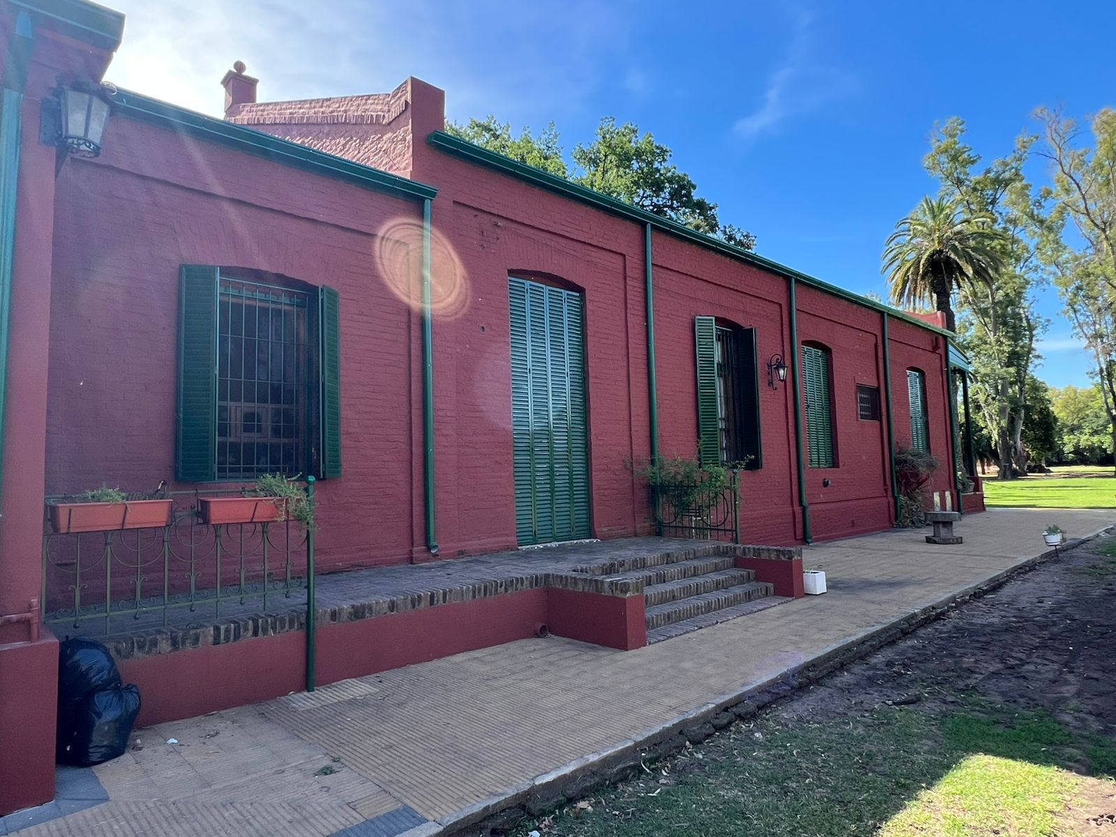 Edificio de ladrillo rojo con contraventanas y puertas verdes junto a un sendero pavimentado y árboles.