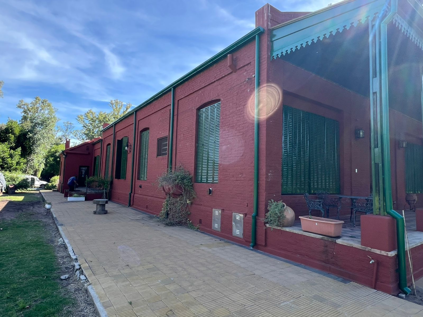 Edificio de ladrillo rojo con contraventanas verdes y un sendero pavimentado bajo un cielo azul soleado.
