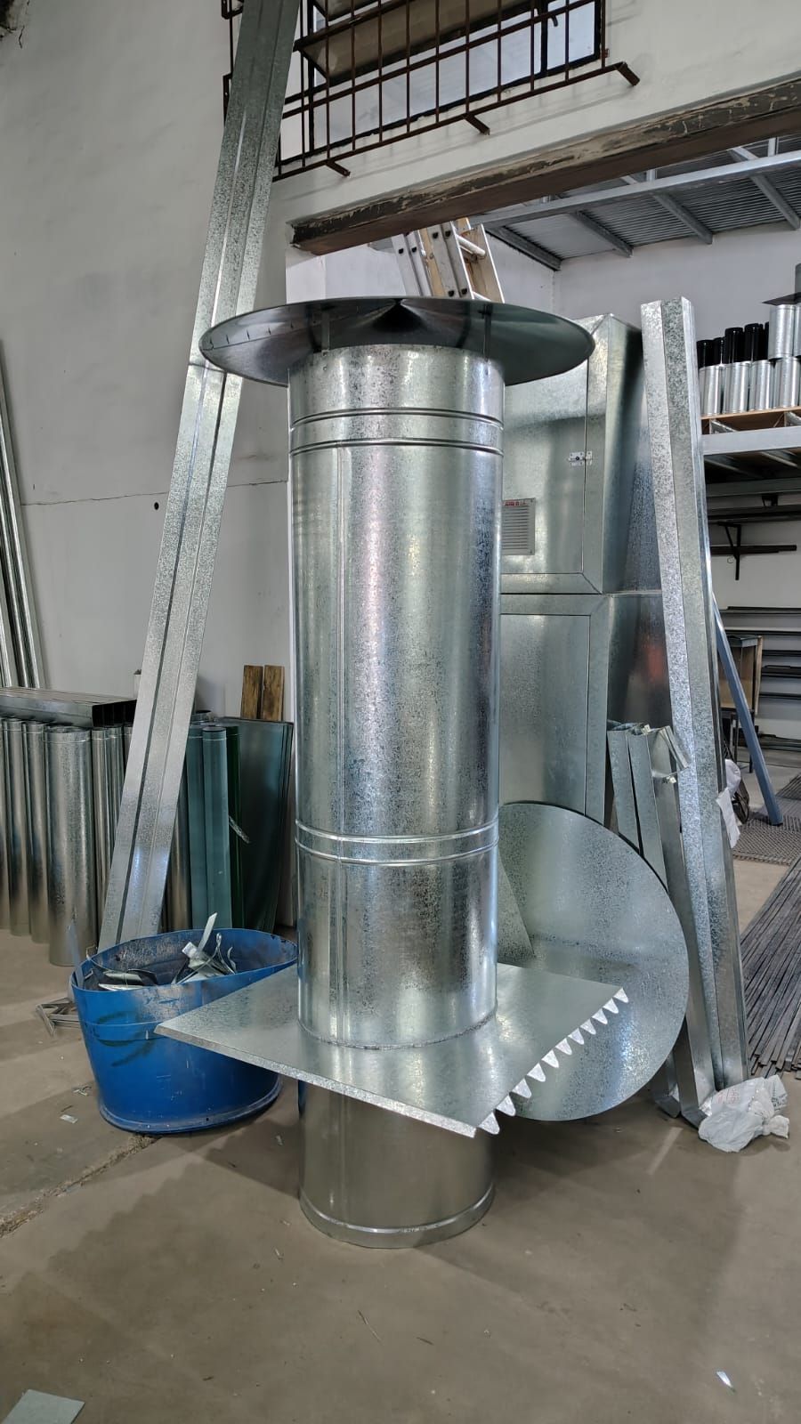 Estufa de chimenea metálica en un taller, con conductos, un cubo azul y mangueras enrolladas a su alrededor.