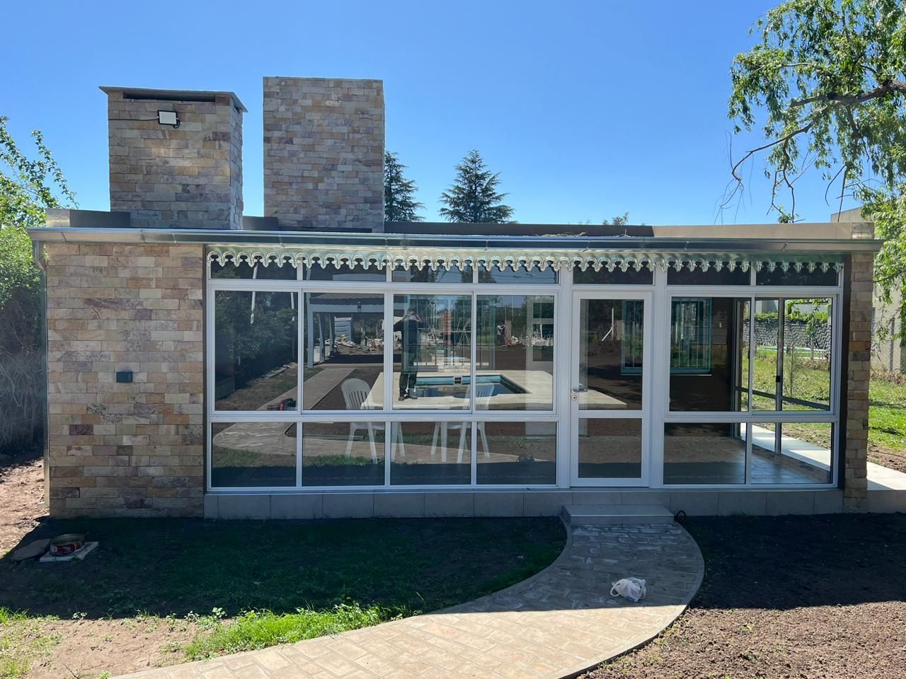Casa moderna de piedra con grandes ventanales y patio en un jardín soleado.