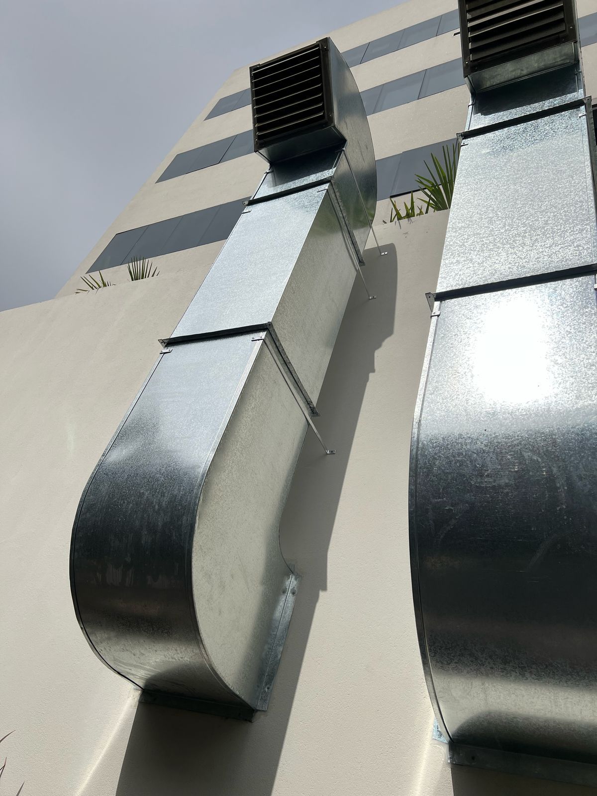 Conductos metálicos curvos y brillantes en la fachada a rayas de un edificio contra un cielo gris.