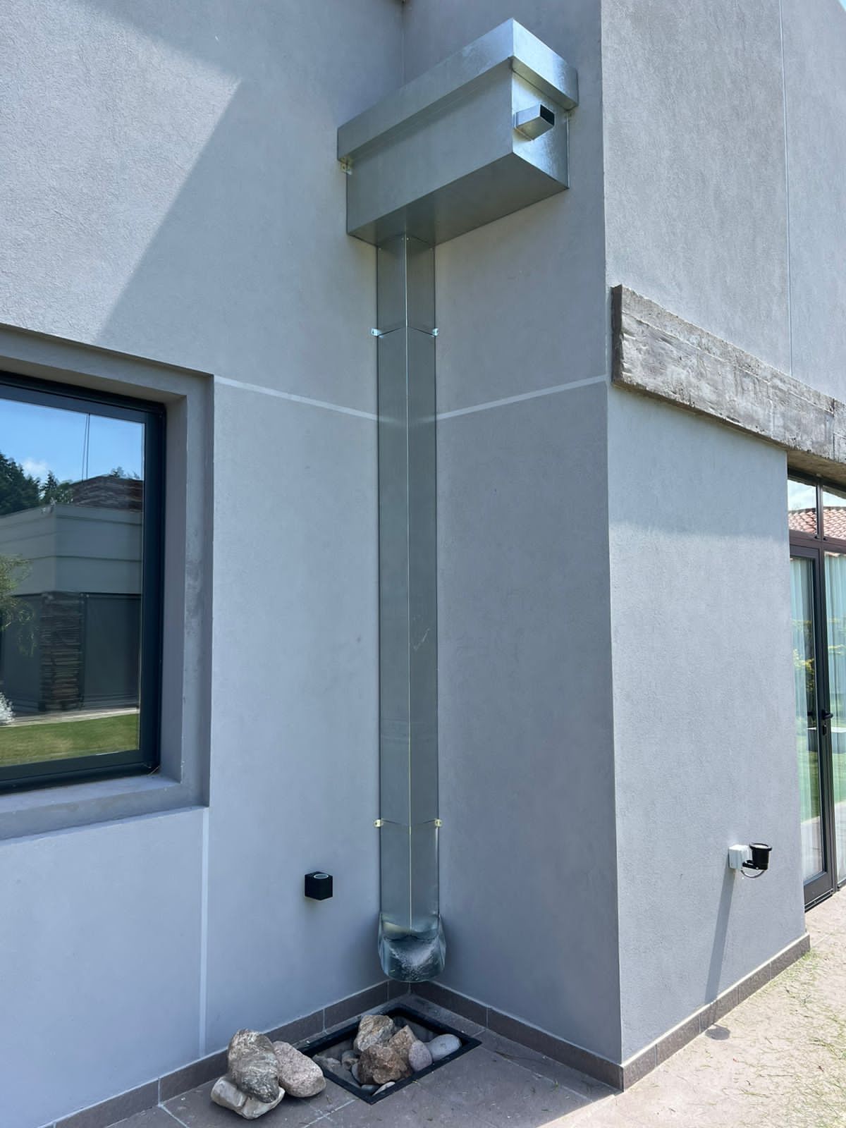 Esquina de un edificio gris con un bajante metálico vertical y una rejilla de ventilación