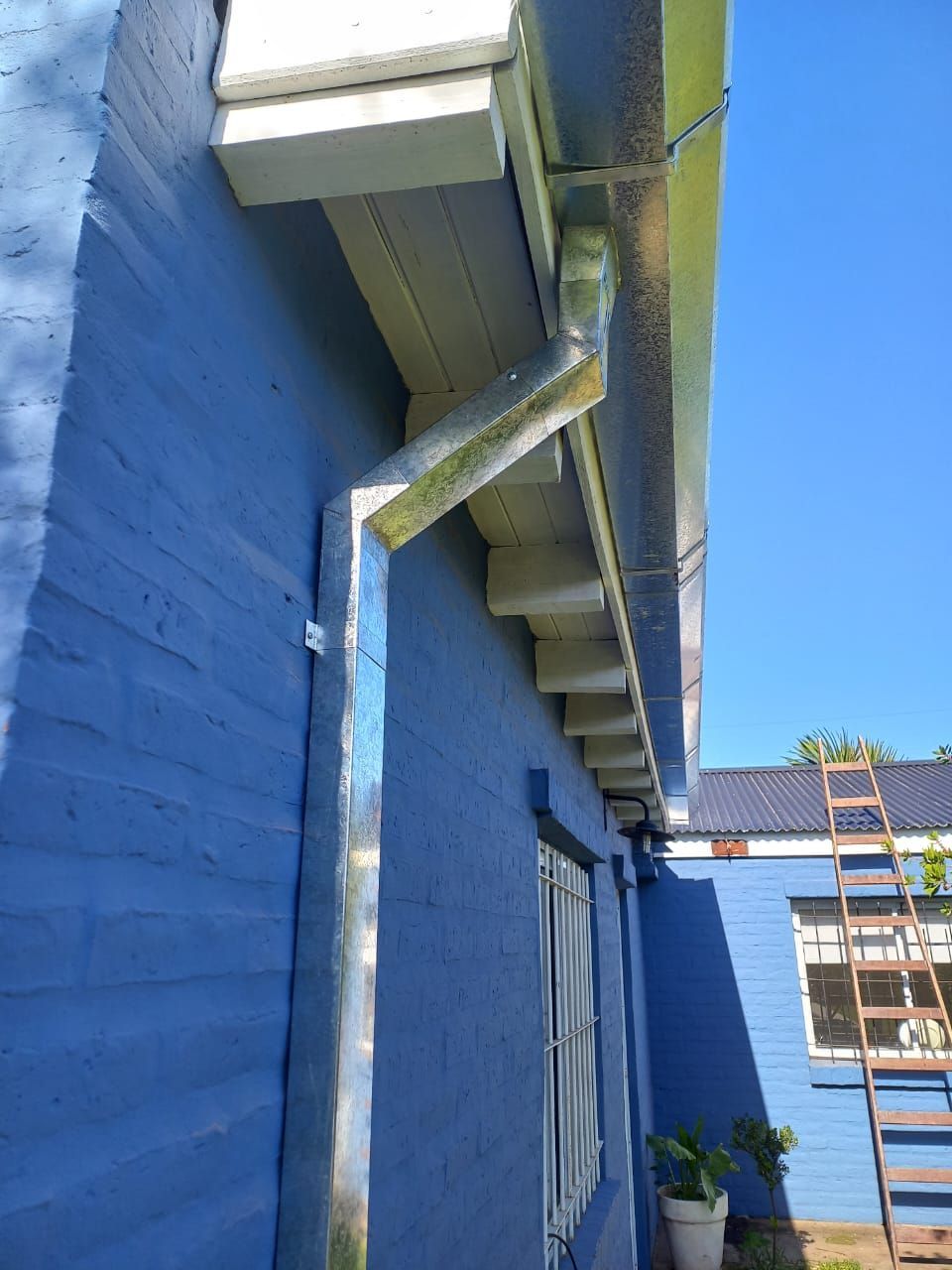 Muro exterior azul con soportes de toldo blancos y una ventana en una zona de patio estrecha.