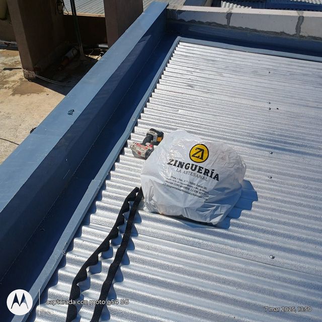 Techo de chapa ondulada blanca con una bolsa de Zinguería y herramientas junto a un muro de parapeto azul.