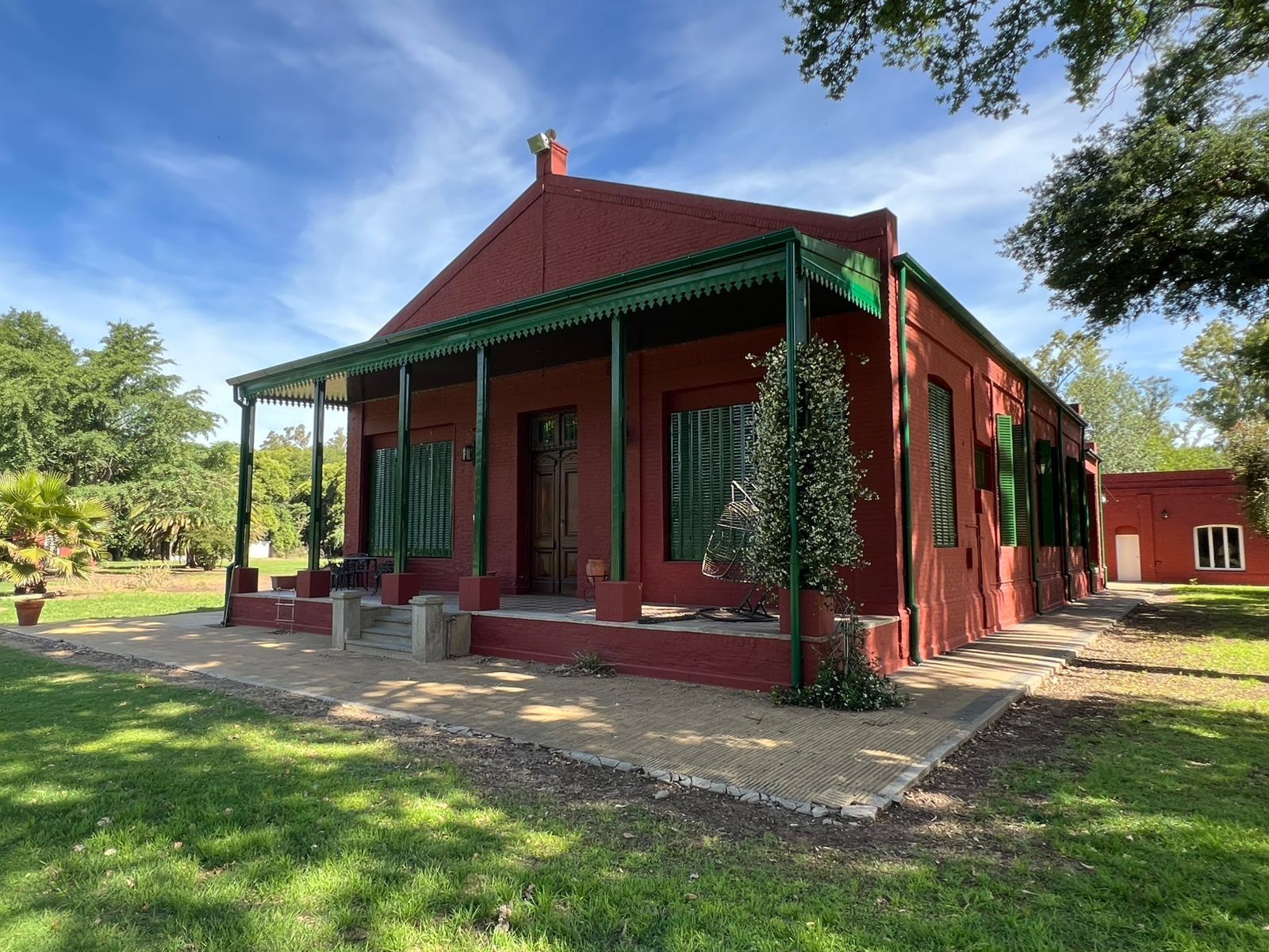 Casa roja con detalles verdes y porche en un soleado jardín con césped, rodeada de árboles.