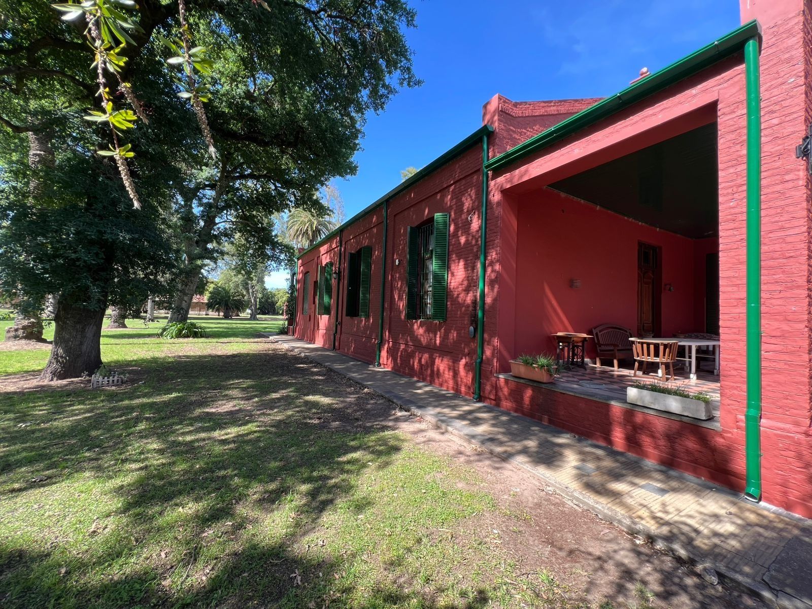 Edificio de ladrillo rojo con detalles verdes y un porche sombreado bajo un gran árbol en un día soleado.