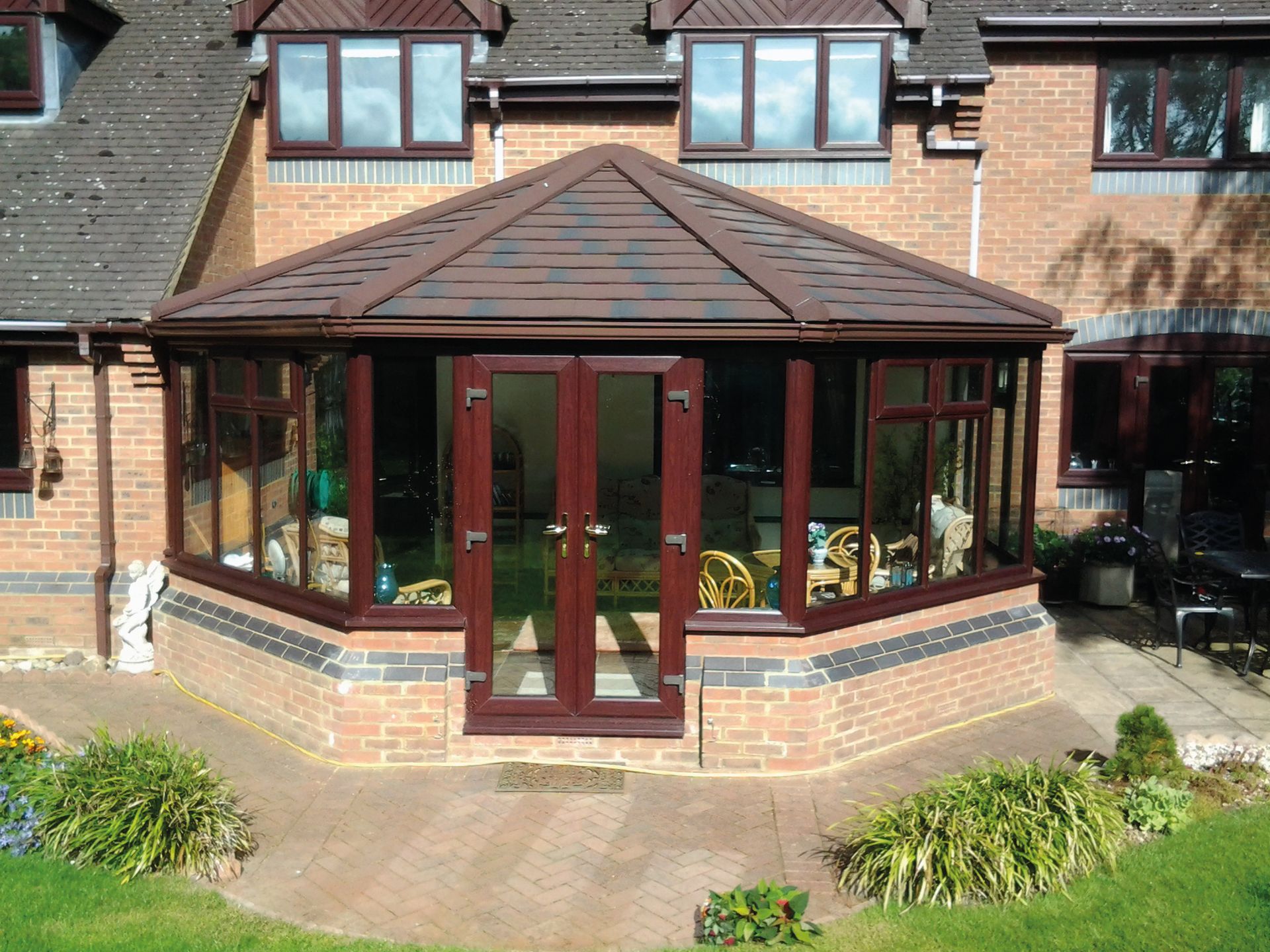 Brown conservatory with a tiled roof, brick base, and surrounding lawn, attached to a two-story brick house.